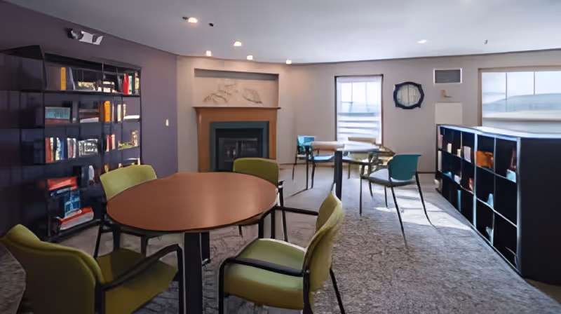 A well-lit common area with round tables and green chairs arranged around them. There are bookshelves filled with books and other items on both sides of the room. A fireplace with a wooden mantel is centered on the back wall between two windows with blinds. The room has a carpeted floor and recessed ceiling lights.