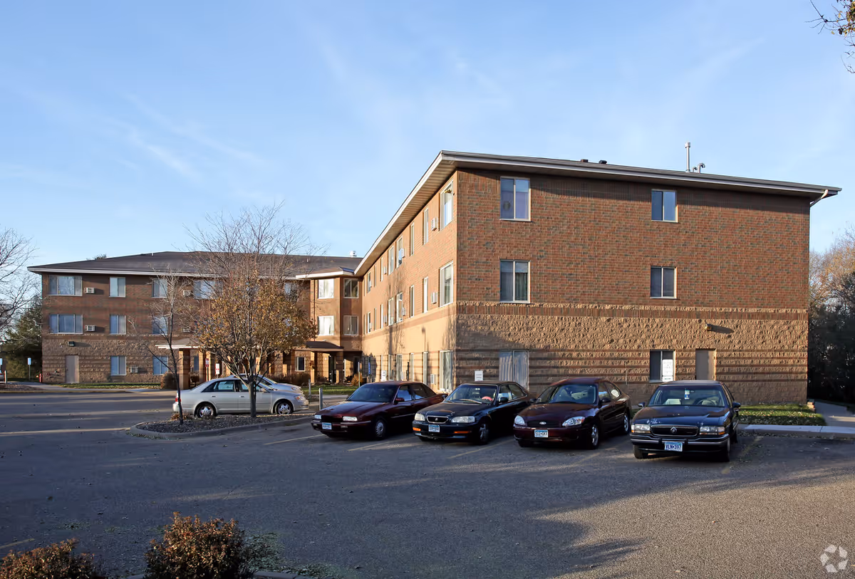 Exterior view of a three-story brick apartment building with several parked cars in front and a small tree in the parking lot under a clear blue sky.