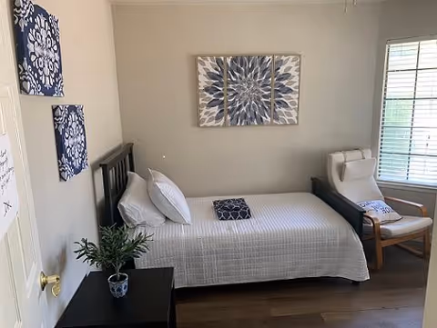 A small bedroom with a single bed covered in a white quilt and two pillows. Above the bed is a three-panel floral artwork in shades of blue and gray. To the left of the bed is a black nightstand with a small potted plant. On the right side of the room is a wooden armchair with white cushions near a window with blinds. The walls are light-colored and the floor is wooden.