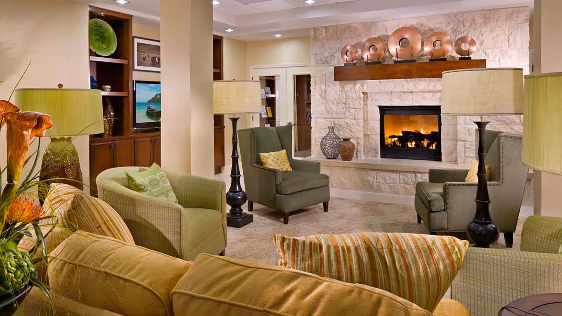 Cozy living room area with a stone fireplace lit with a warm fire. The room features green and beige armchairs, a beige sofa with striped pillows, two tall black floor lamps with beige lampshades, and decorative items on shelves and mantel. The walls are light-colored, and the room has a comfortable and inviting atmosphere.