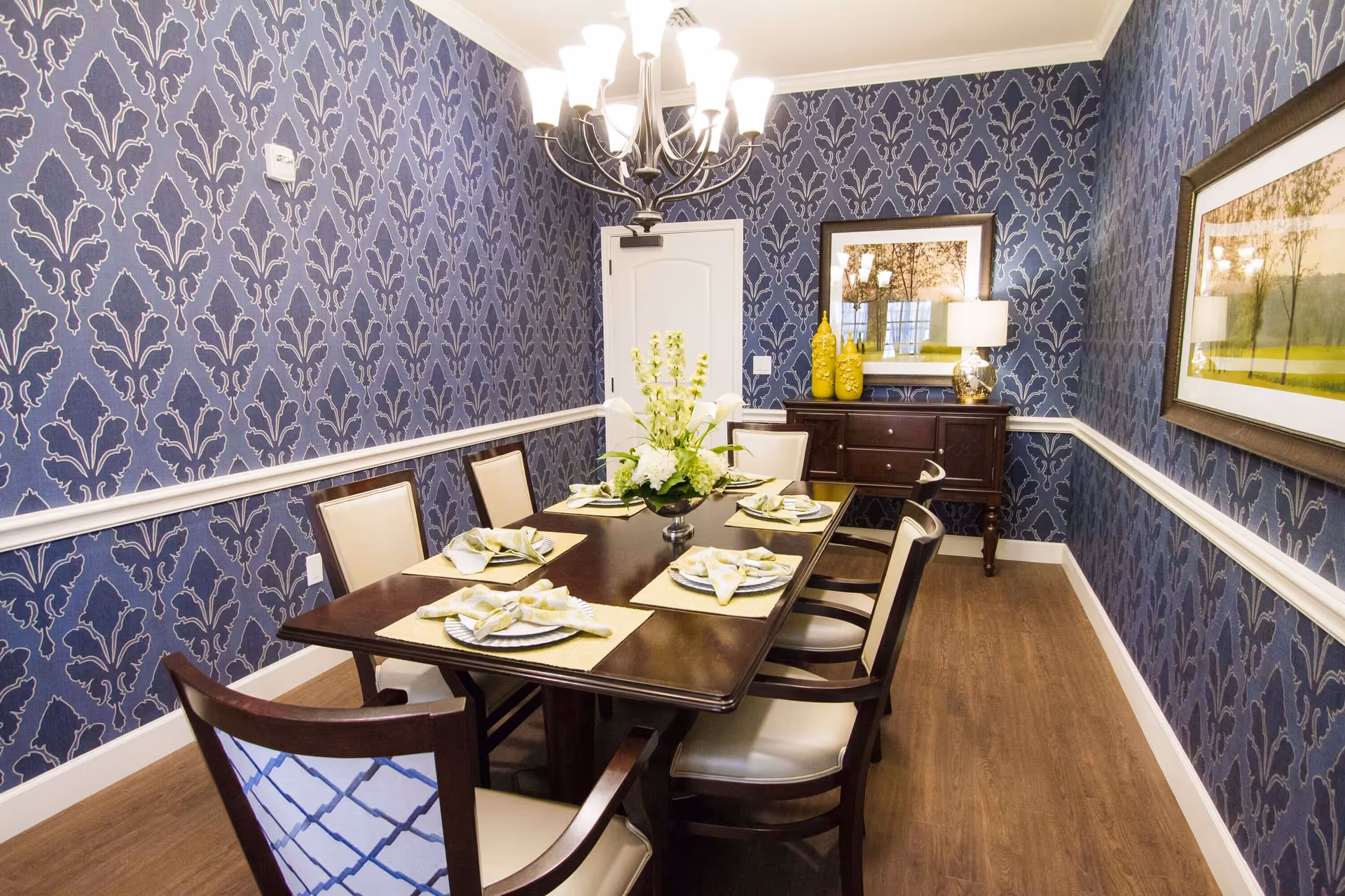 A dining room with a dark wooden table set for six people with beige cushioned chairs. The walls are covered in blue patterned wallpaper with white trim. A chandelier with multiple lights hangs above the table. There is a sideboard against the far wall with decorative yellow vases and a lamp, and two framed landscape pictures hang on the walls.
