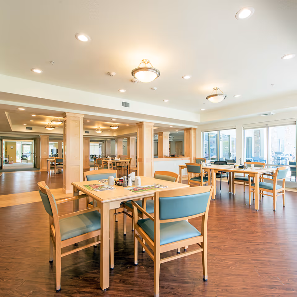 A bright and spacious dining room with wooden tables and chairs featuring teal cushions. The room has large windows allowing natural light to fill the space, wooden flooring, and ceiling lights. The tables are set with placemats, salt and pepper shakers, and napkin holders.