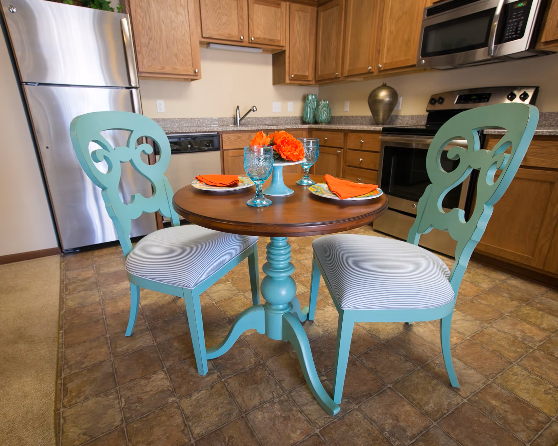 A small kitchen dining area with a round wooden table and two light blue chairs with striped cushions. The table is set with two plates, orange napkins, two blue glasses, and a centerpiece with orange flowers. The kitchen has wooden cabinets, a stainless steel refrigerator, stove, microwave, and a dishwasher.
