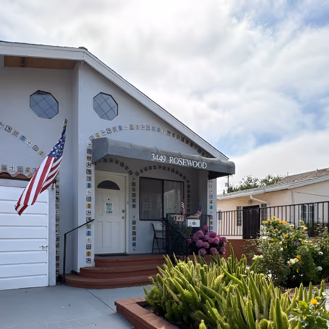 Front entrance of a light-gray house with an awning reading "3449 ROSEWOOD", steps, an American flag, and a small flowering garden.
