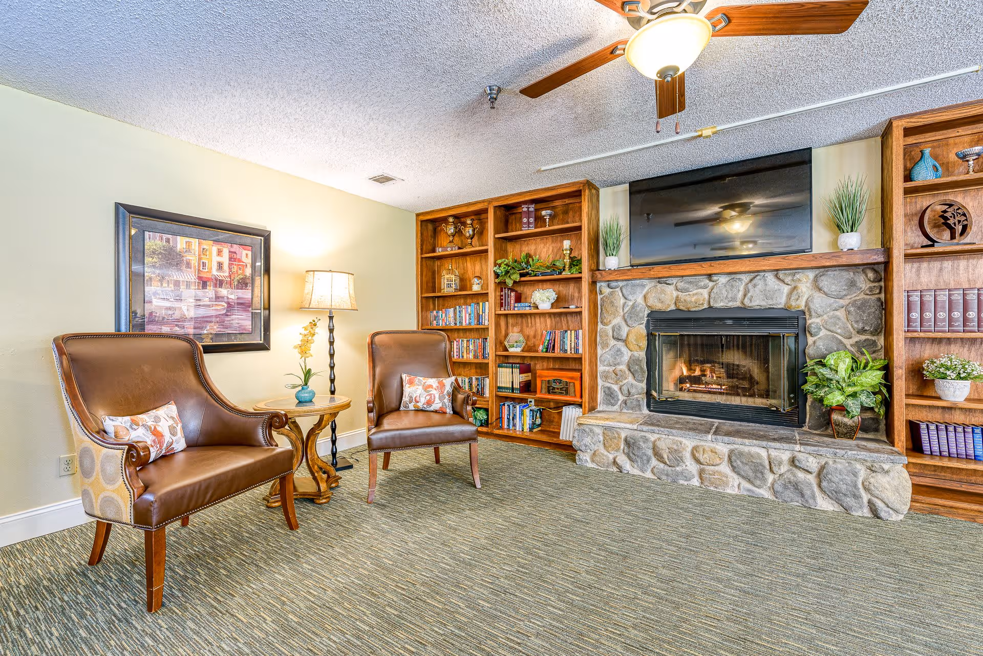 A cozy living room area with two brown leather armchairs, a small round wooden table with a lamp and a flower vase, a stone fireplace with a fire burning, a flat-screen TV mounted above the fireplace, and wooden bookshelves filled with books and decorative items on either side of the fireplace.