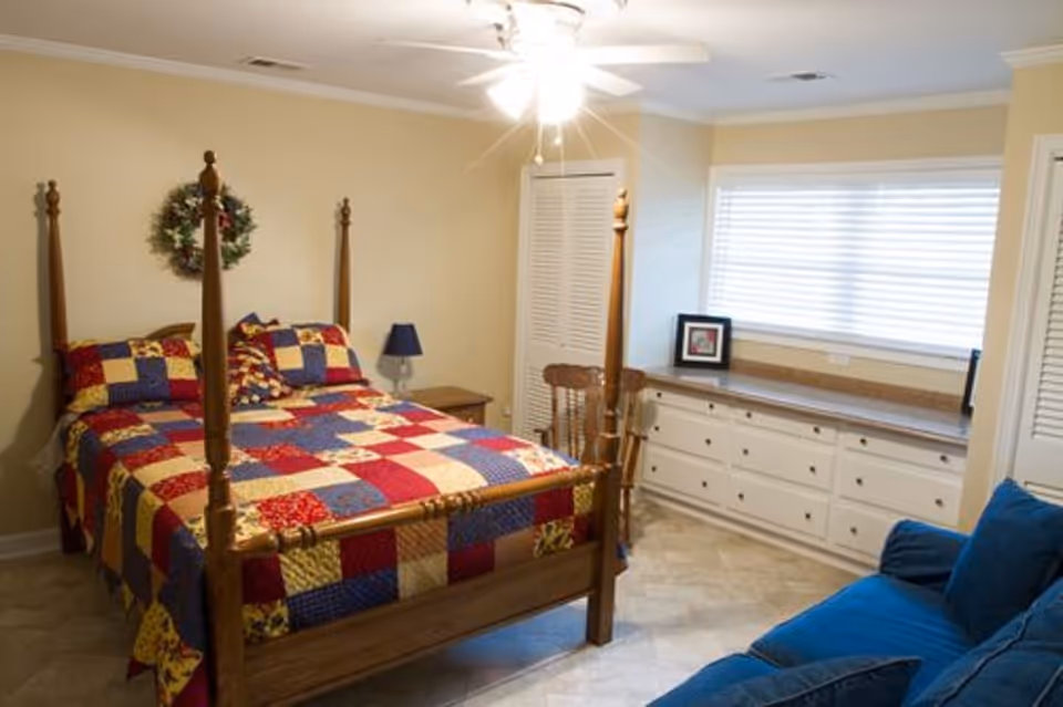 A cozy bedroom with a wooden four-poster bed covered with a colorful patchwork quilt. There is a small wooden nightstand with a blue lamp beside the bed. A wooden chair is placed near a long built-in dresser with multiple drawers under a large window with blinds. A blue sofa is partially visible in the foreground. The walls are painted light beige and the floor has a light-colored tile.