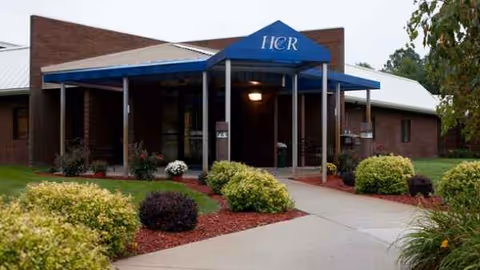 Front entrance of a brick senior living facility with a blue canopy and landscaped walkway