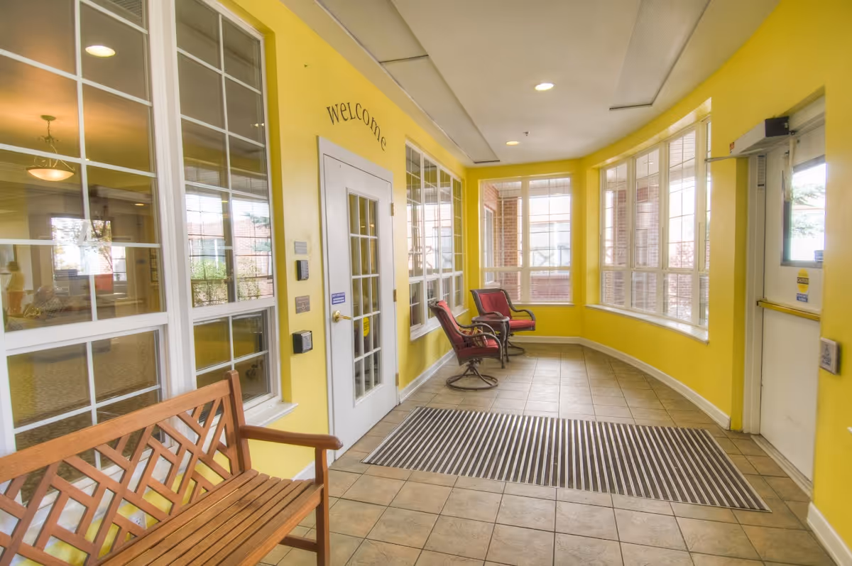 A bright indoor seating area with yellow walls and large windows. There is a wooden bench on the left and two red cushioned chairs on the right near a white door with a 'welcome' sign above it. The floor is tiled and there is a striped floor mat near the door on the right side.