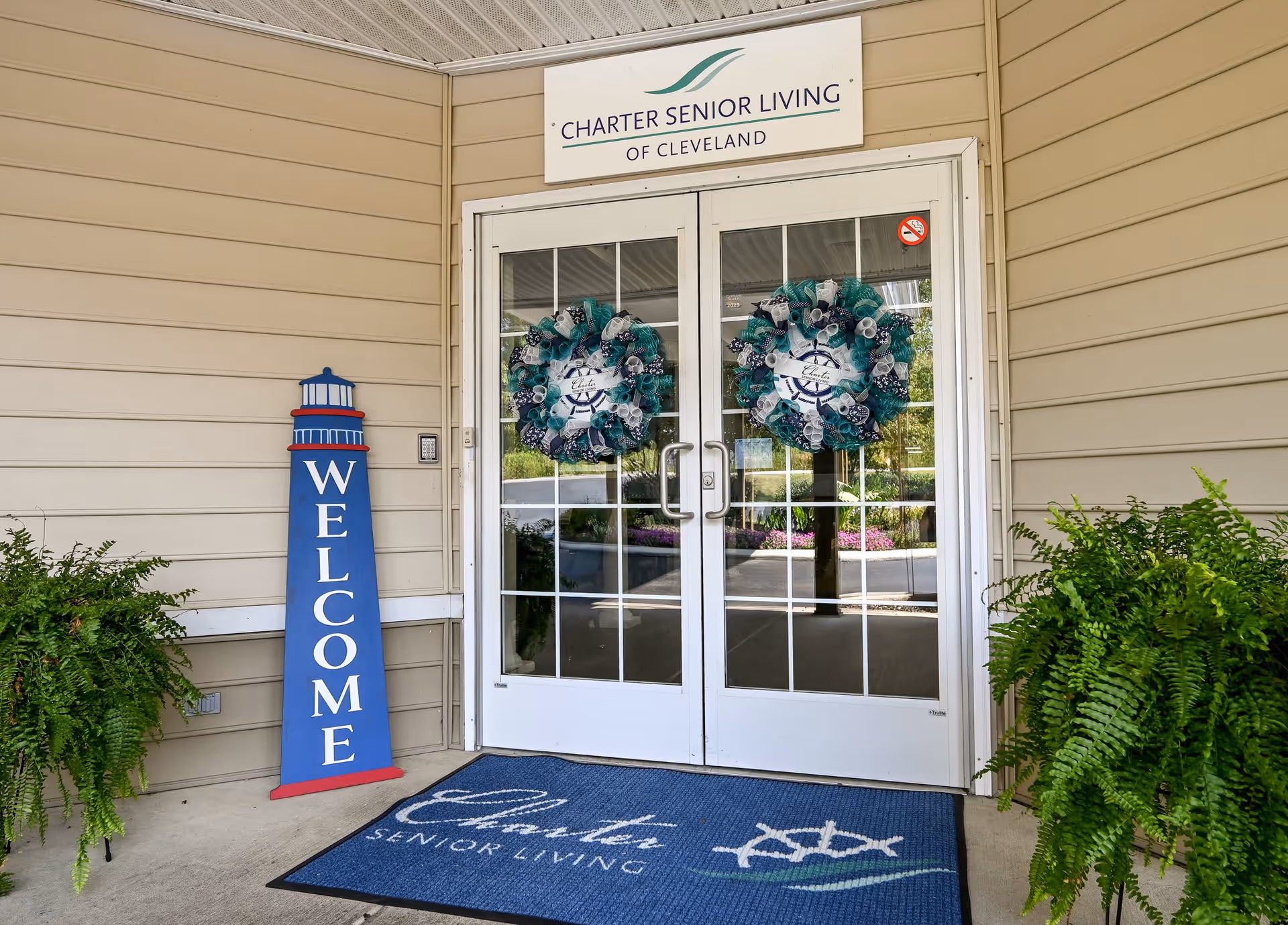 Entrance to Charter Senior Living of Cleveland featuring double glass doors with decorative wreaths, a blue welcome sign shaped like a lighthouse, two large green potted ferns on either side, and a blue welcome mat with the facility's name and logo.