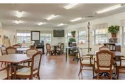 Bright communal dining/activity room with several round tables and chairs, hardwood floors, large windows, plants, and a wall-mounted TV.