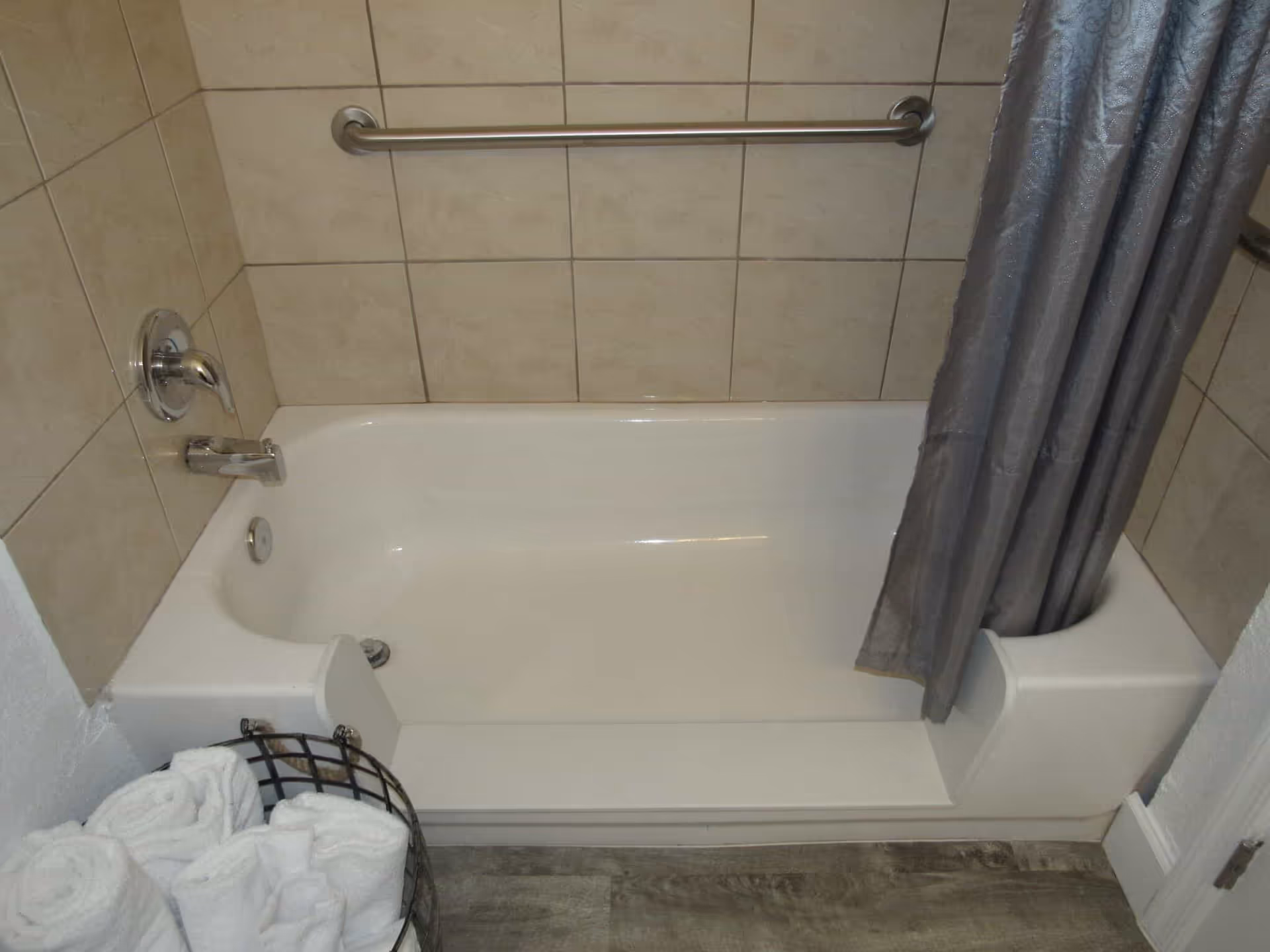 White bathtub with tiled walls, stainless grab bar and faucet, gray shower curtain, and a basket of rolled towels on the floor.