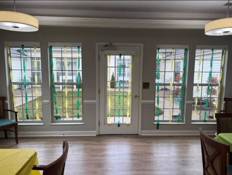 Interior view of a room with five tall windows and a glass door in the center, decorated with hanging green and yellow streamers. The room has light wood flooring, a round ceiling light fixture, and tables with yellow and green tablecloths partially visible on the sides.