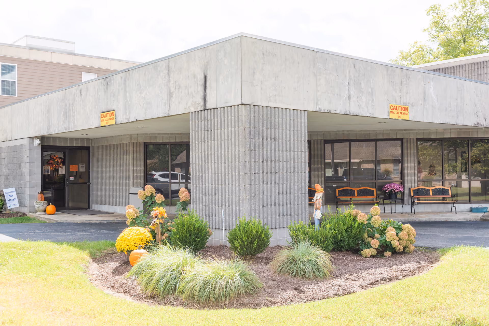 Exterior view of a senior living facility entrance with a concrete overhang supported by a large pillar. There are autumn decorations including pumpkins and flowers near the entrance. Two benches and a small table with a flower pot are visible under the overhang. A sign near the door reads 'Support Our Heroes'.