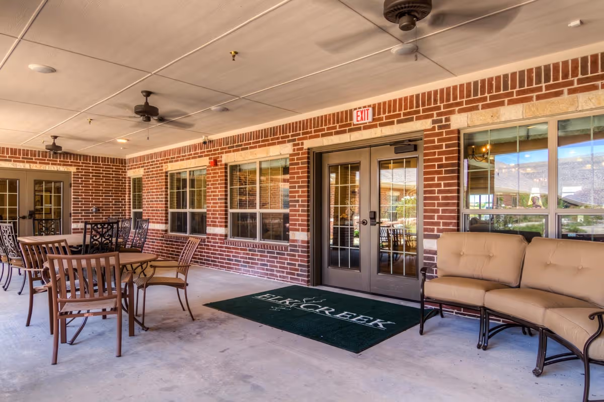 Covered outdoor patio area at Elk Creek Assisted Living and Memory Care with brick walls, ceiling fans, multiple tables and chairs, and a cushioned bench. A green floor mat with the Elk Creek logo is placed in front of double glass doors.