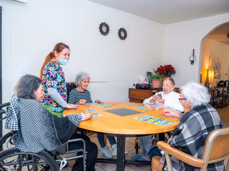 A group of four elderly women sitting around a round wooden table playing a card game, accompanied by a caregiver wearing a colorful floral scrub top and a face mask. The room has white walls with two decorative mirrors and a wooden cabinet with a potted plant and a radio in the background.