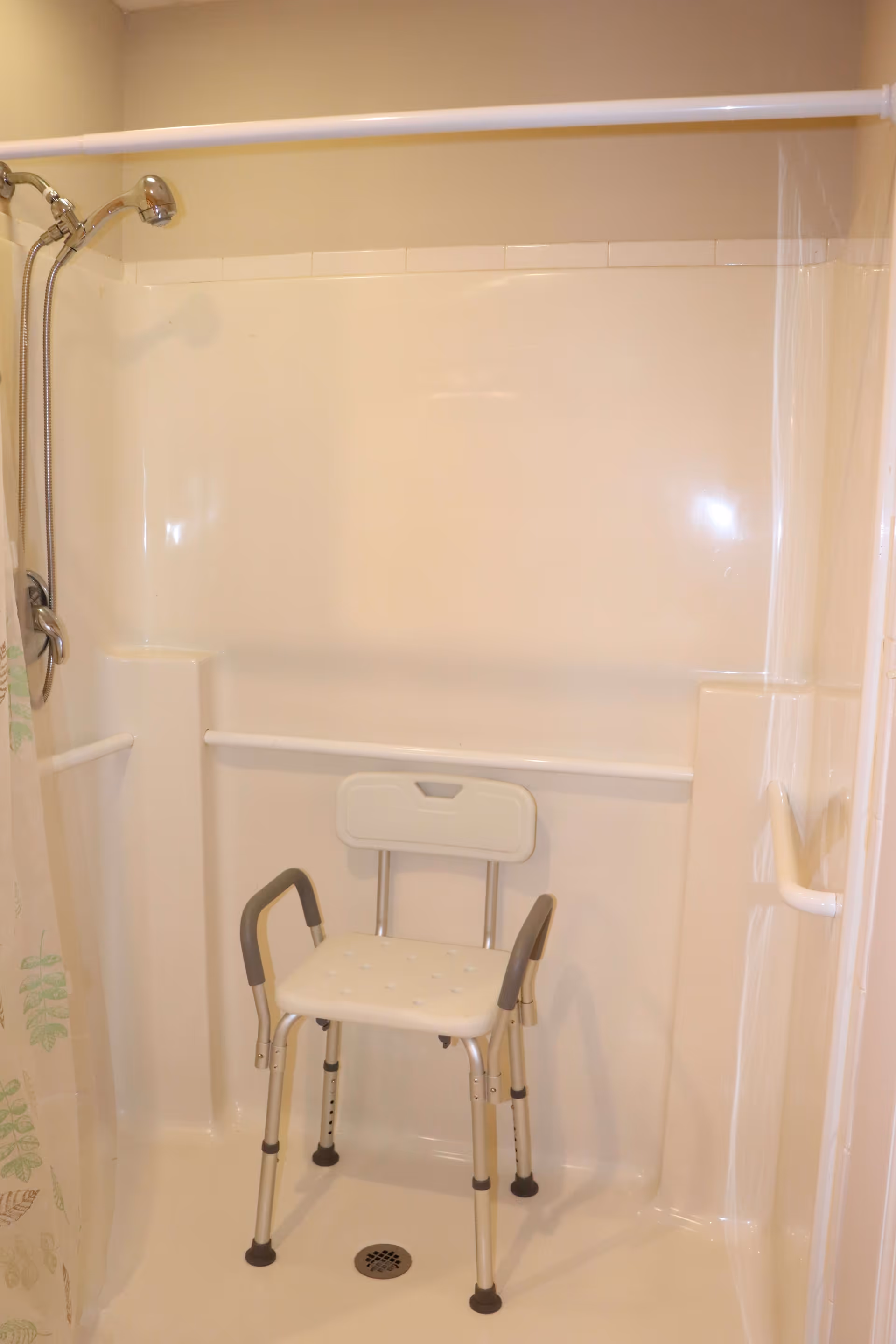 A white shower stall with a handheld showerhead mounted on the left wall, a white shower curtain with leaf patterns partially visible on the left, and a white shower chair with armrests and a backrest positioned inside the shower.
