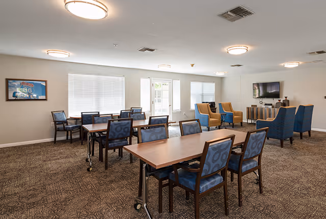 A spacious senior living common area with multiple tables and chairs arranged for group seating. The room has carpeted floors, several windows with blinds, and ceiling lights. There is a seating area with blue and yellow armchairs near a wall-mounted television.