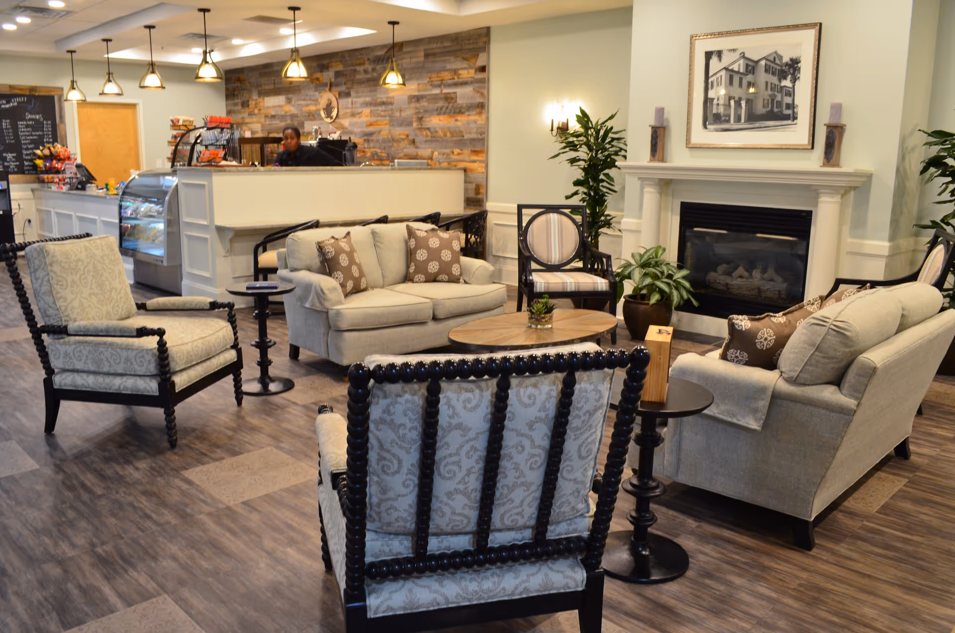 A cozy lounge area with beige sofas and patterned armchairs arranged around a wooden coffee table. There is a fireplace with a framed picture above it and potted plants on either side. In the background, a counter with a staff member behind it and a display case with snacks and treats is visible, illuminated by hanging pendant lights.
