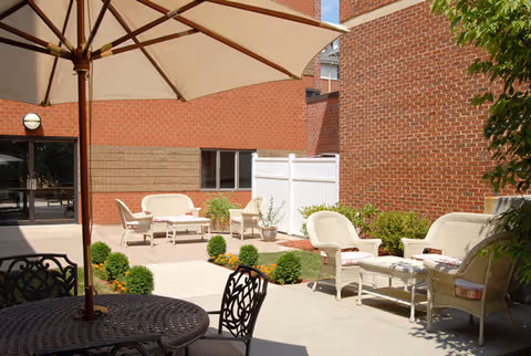 Outdoor patio area with wicker chairs and a table under a large beige umbrella, surrounded by brick walls and some greenery including small bushes and potted plants.