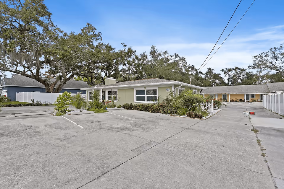 Single-story building with a paved parking lot, landscaped greenery, and large oak trees under a blue sky.