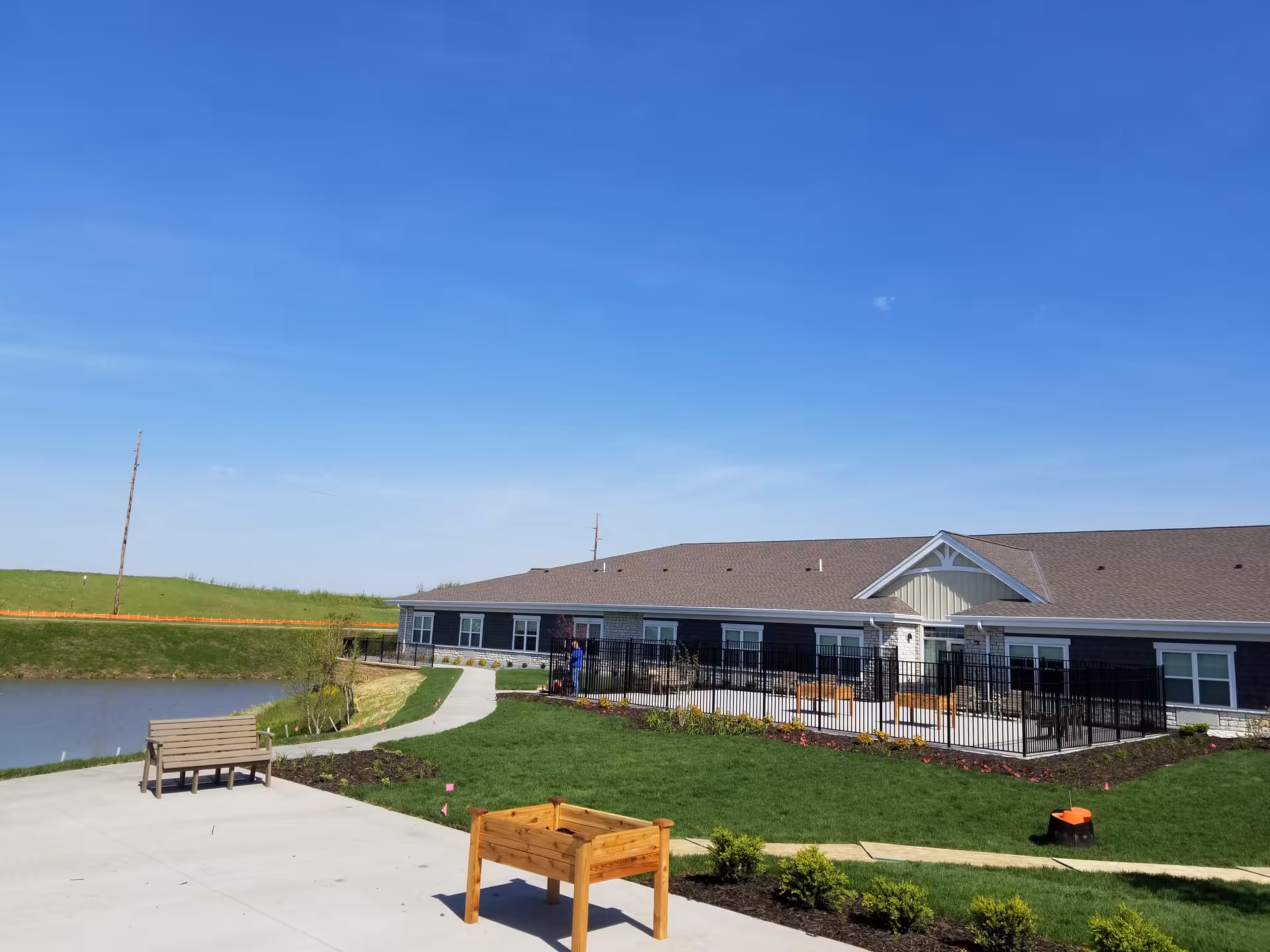 Single-story senior living building with a fenced outdoor patio, lawn, pond, benches, and a wooden planter under a clear blue sky.