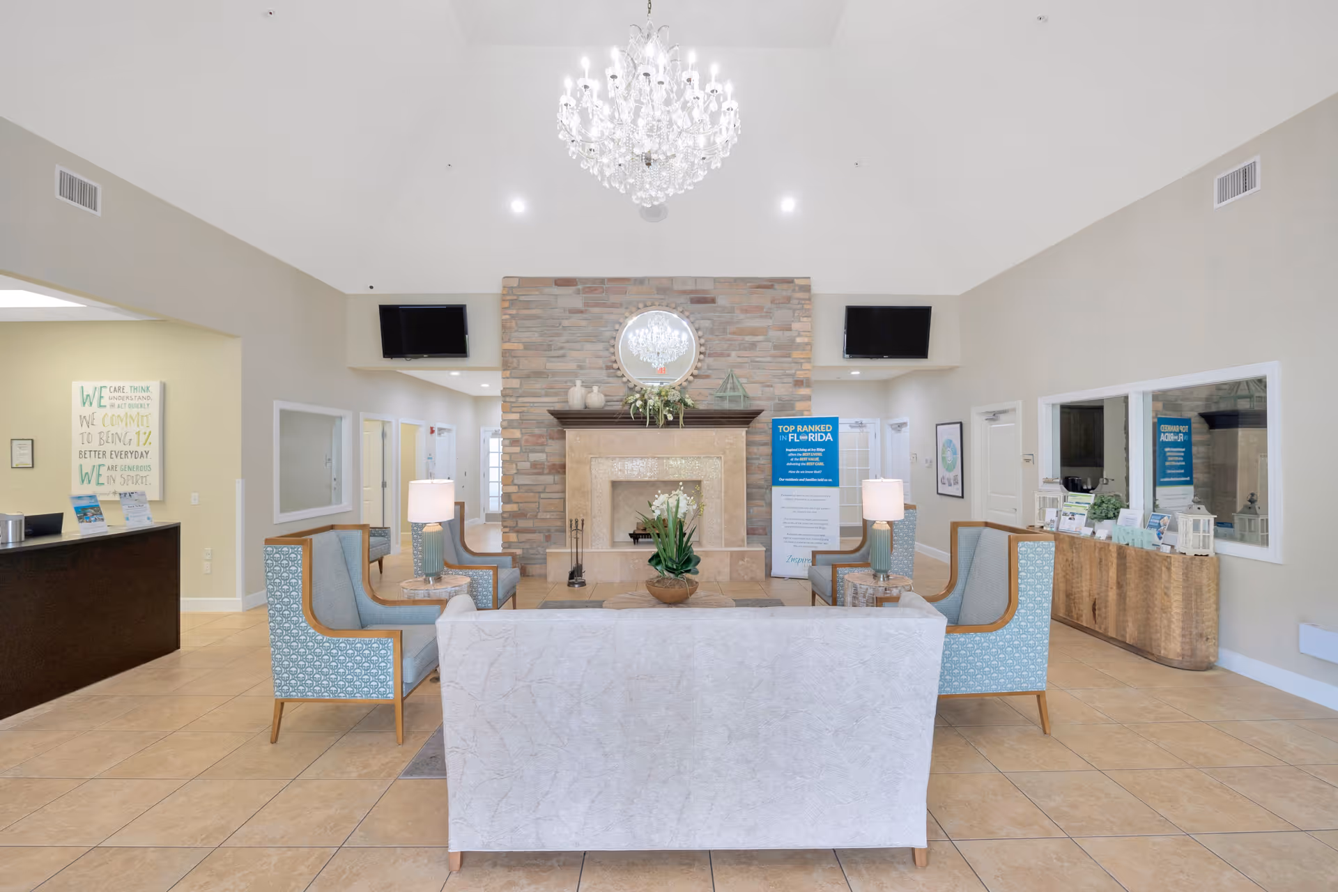 Bright and spacious senior living facility common area with a stone fireplace, a round mirror above it, and a chandelier hanging from the ceiling. The room features a white sofa and four blue patterned armchairs arranged around a central coffee table with a plant. There are two flat-screen TVs mounted on the walls on either side of the fireplace, lamps on side tables, and informational signs near the fireplace. The floor is tiled, and the walls are painted in neutral tones.