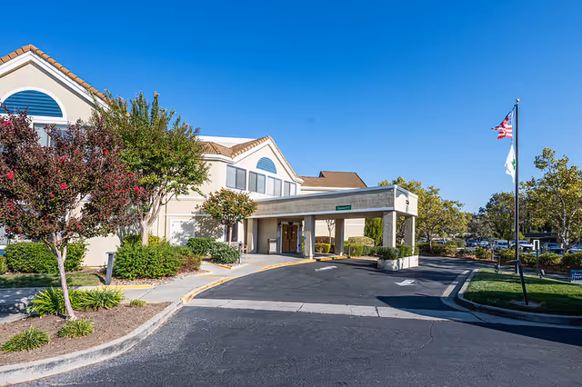 Front entrance of a post-acute facility showing a covered drop-off canopy, landscaped grounds, and flagpoles under a clear blue sky.