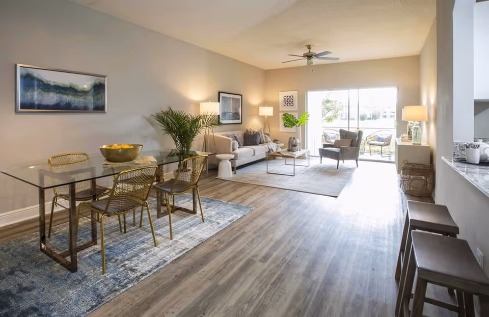 A bright and spacious living and dining area with a glass dining table and four gold wire chairs on a blue rug. The living room features a beige sofa, an armchair, a coffee table, floor lamps, and a ceiling fan. Large sliding glass doors open to an outdoor seating area with two chairs and a small table.