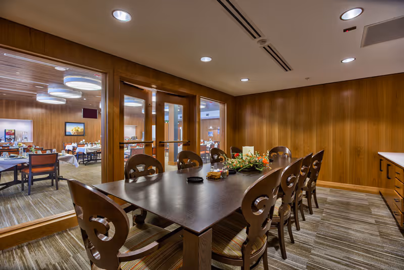 A dining room with a long dark wooden table surrounded by eight matching chairs with decorative backs. The table has a floral centerpiece and some small items on it. The room has wood-paneled walls and carpeted floors. Through large glass windows, another dining area with tables and chairs is visible, featuring round ceiling lights and a framed picture on the far wall.