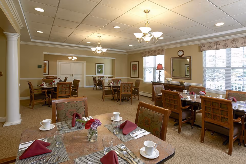 A well-lit dining room with multiple tables set for a meal, featuring maroon napkins, white cups, glasses, and silverware. The room has beige walls, carpeted floors, large windows with patterned valances, framed artwork, and chandeliers hanging from the ceiling. There are wooden chairs with cushioned seats around the tables.