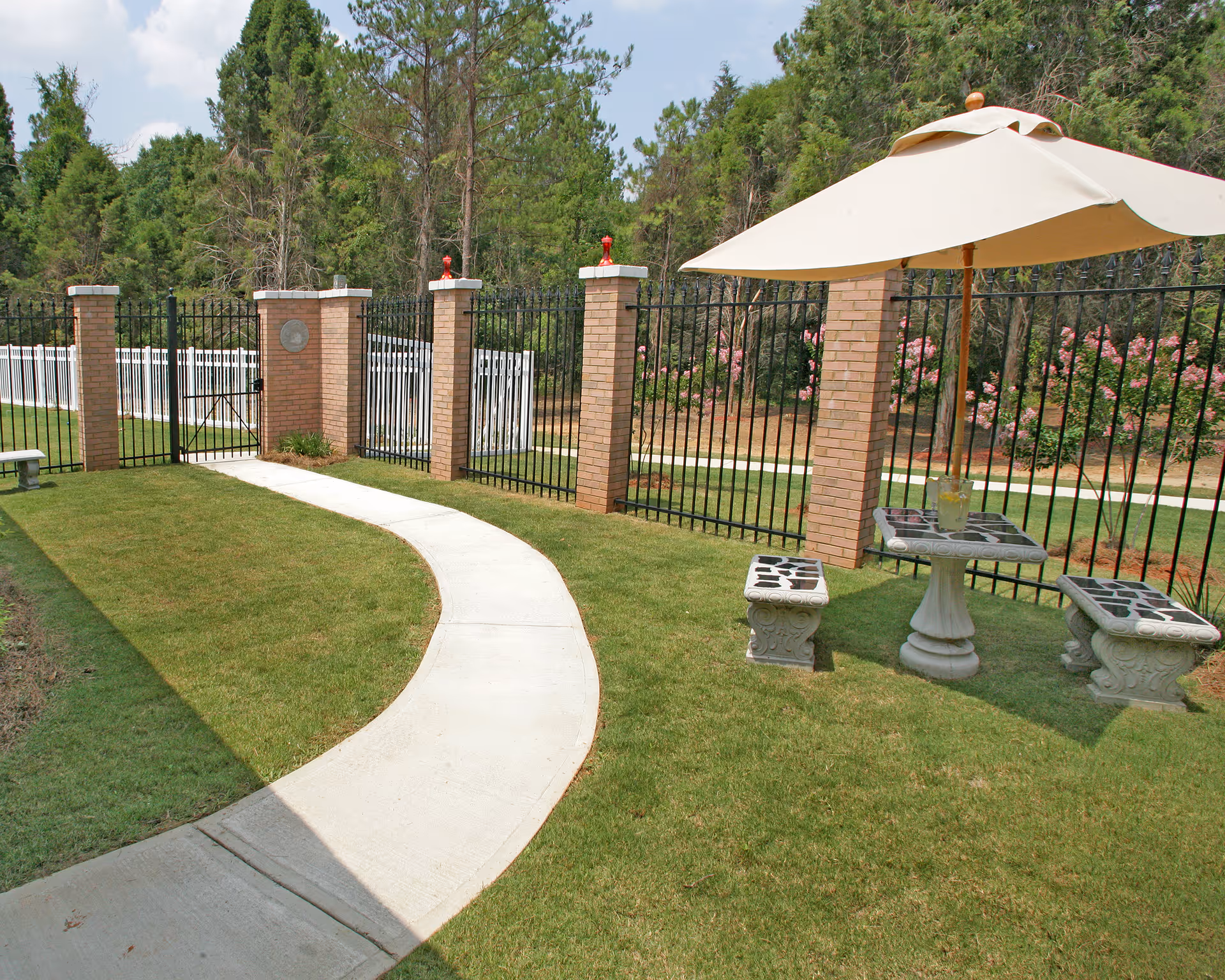A fenced outdoor garden area with a curved concrete pathway, green grass, and a small seating arrangement consisting of a stone table and two stone benches under a large beige umbrella. Trees and flowering bushes are visible beyond the fence.