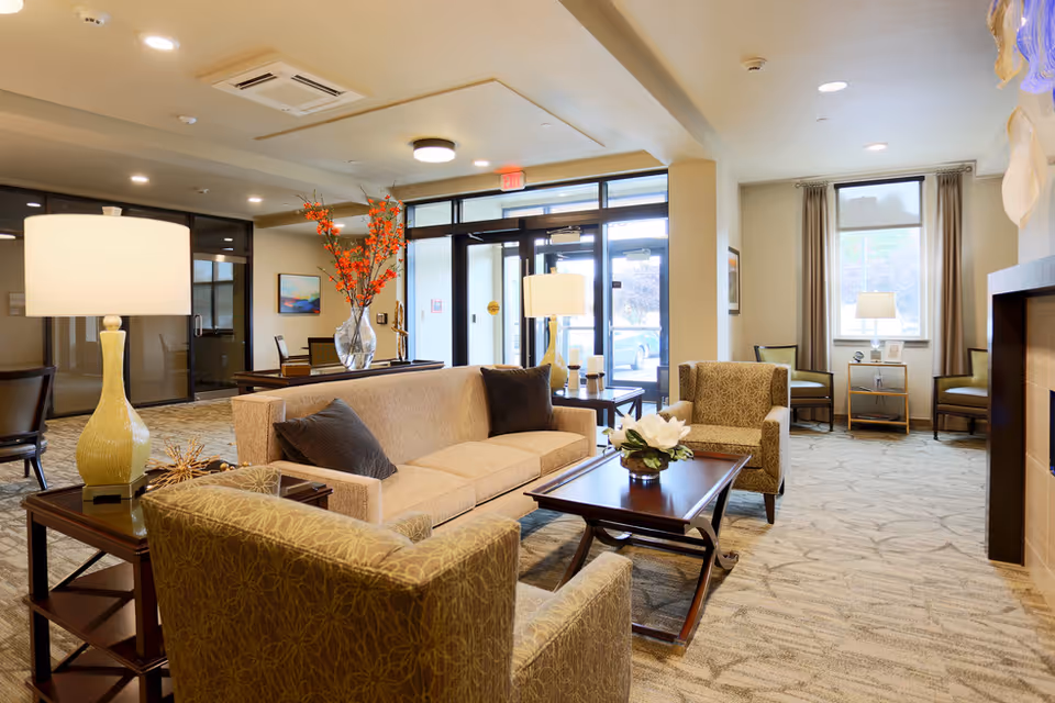 A well-lit senior living facility lounge area with a beige sofa, two patterned armchairs, a wooden coffee table with a flower arrangement, and side tables with lamps. Large windows and glass doors allow natural light to enter, and there are decorative elements such as a vase with orange flowers and framed artwork on the walls.