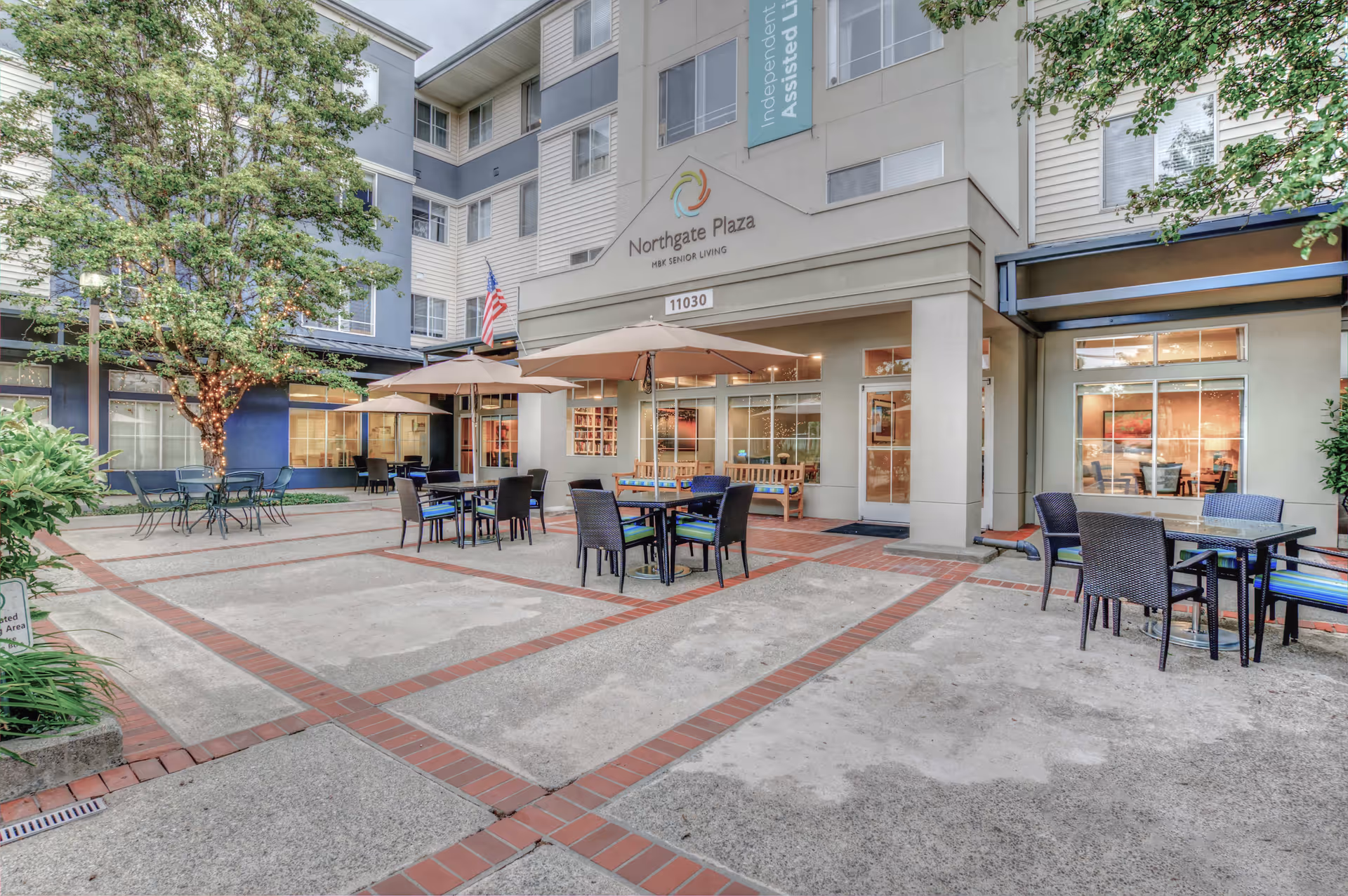 Outdoor courtyard area of Northgate Plaza senior living facility with multiple tables and chairs, some shaded by umbrellas, surrounded by building walls and windows. A tree with string lights is visible on the left side.