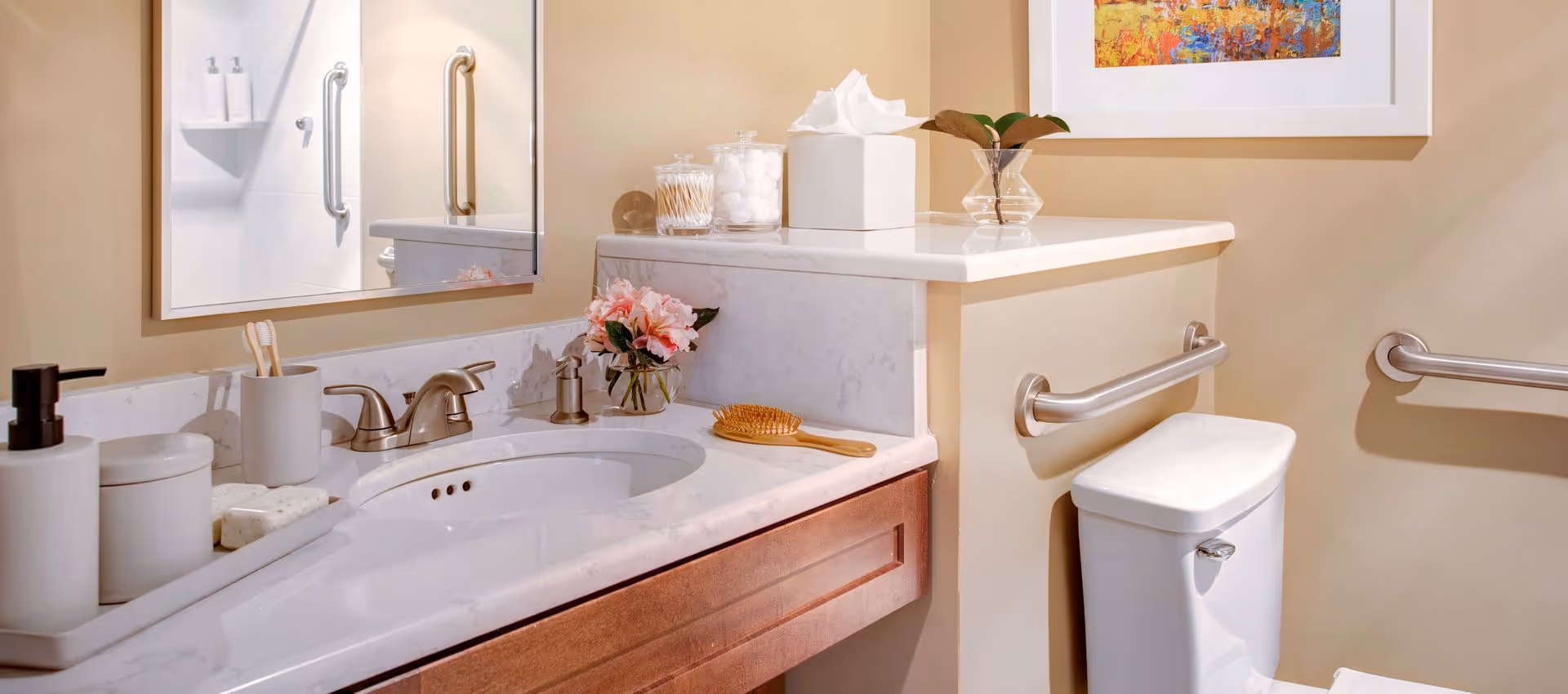 Accessible bathroom with a marble-top sink, mirror, toilet, and stainless steel grab bars, decorated with toiletries and flowers.