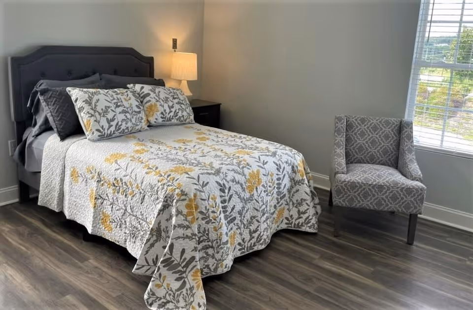 A bedroom with a bed featuring a floral patterned quilt and pillows in shades of gray and yellow. There is a dark upholstered headboard, a nightstand with a lamp, a patterned gray armchair, and a window with white blinds letting in natural light. The floor is wood with a grayish tone.