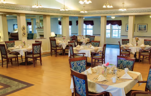 A dining room with multiple tables set with white tablecloths, napkins, glassware, and silverware. The room features wooden chairs with patterned upholstery, large windows with dark valances, and a mirrored wall reflecting the space. The lighting includes ceiling fixtures with multiple bulbs.