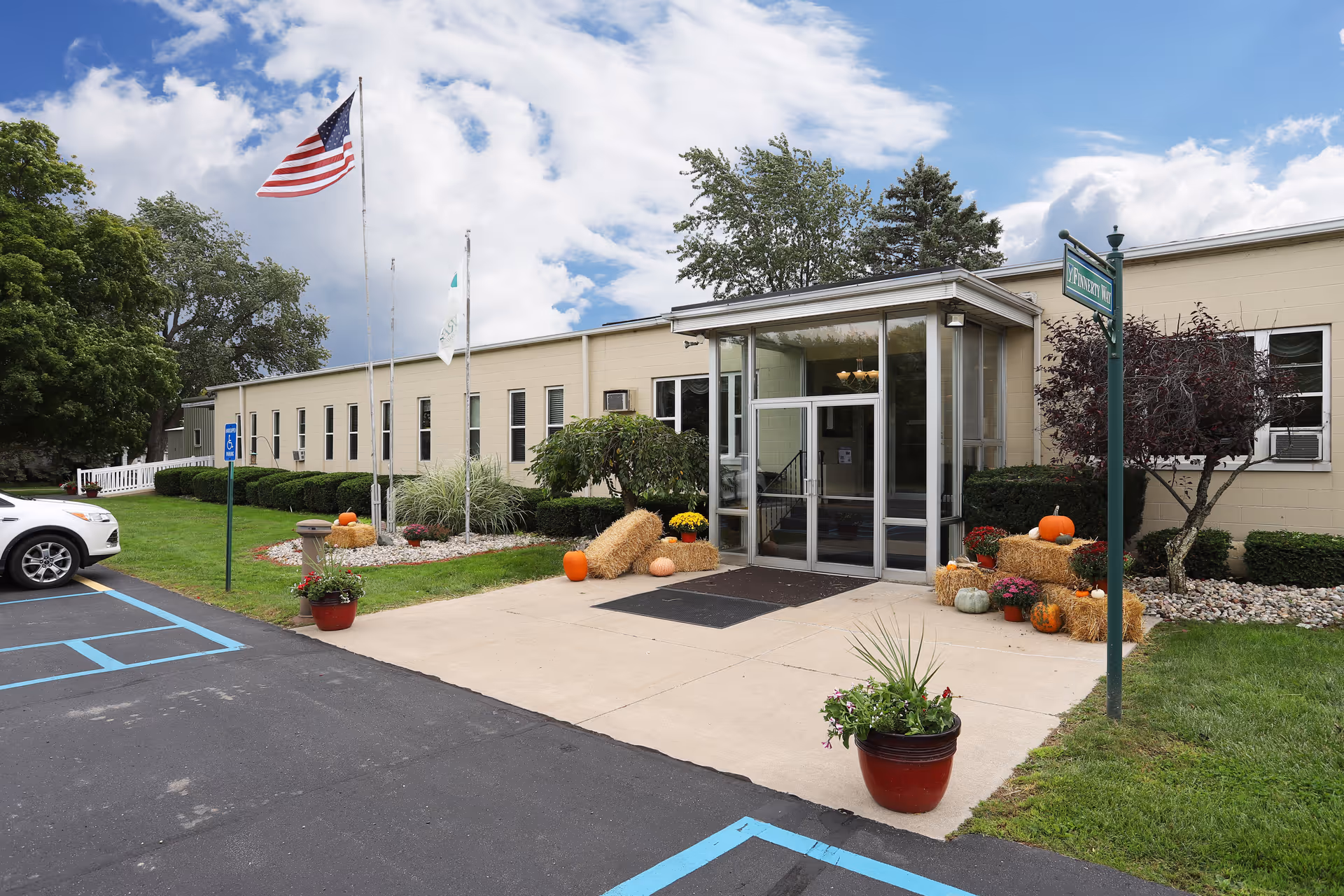 Front entrance of a single-story beige building with a glass vestibule, American flag, pumpkins and hay bale fall decorations, and adjacent parking.