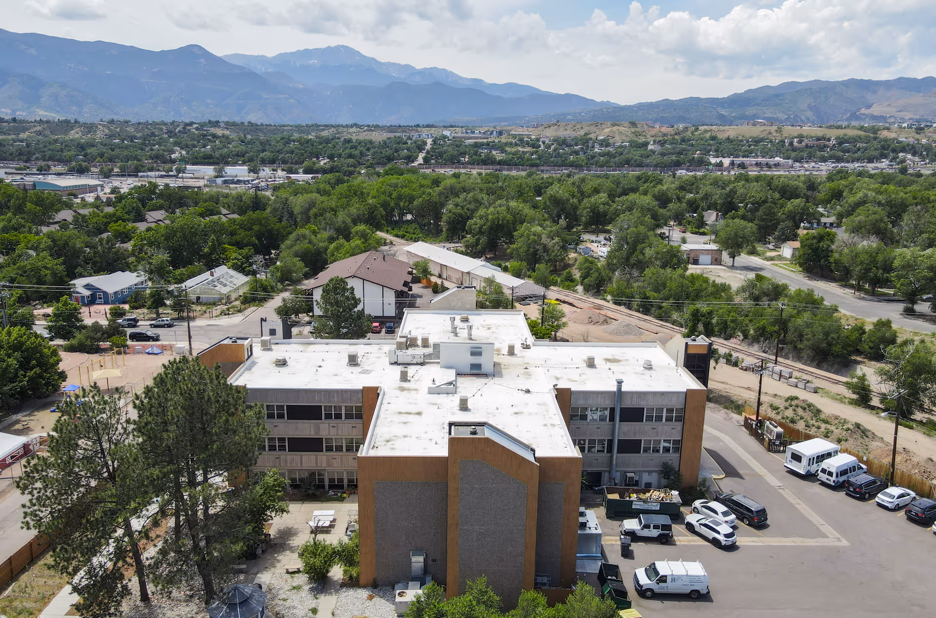 Aerial view of Springs Village Care Center, a multi-story building with a white roof surrounded by trees and parking areas. Several vehicles are parked around the building, with mountains visible in the background under a partly cloudy sky.