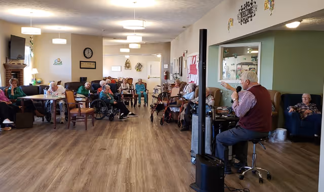 A group of elderly people seated in a spacious room with wooden flooring, some in wheelchairs and others in chairs, attentively listening to a man speaking into a microphone. The room has light-colored walls, ceiling lights, a clock, and some decorations on the walls.