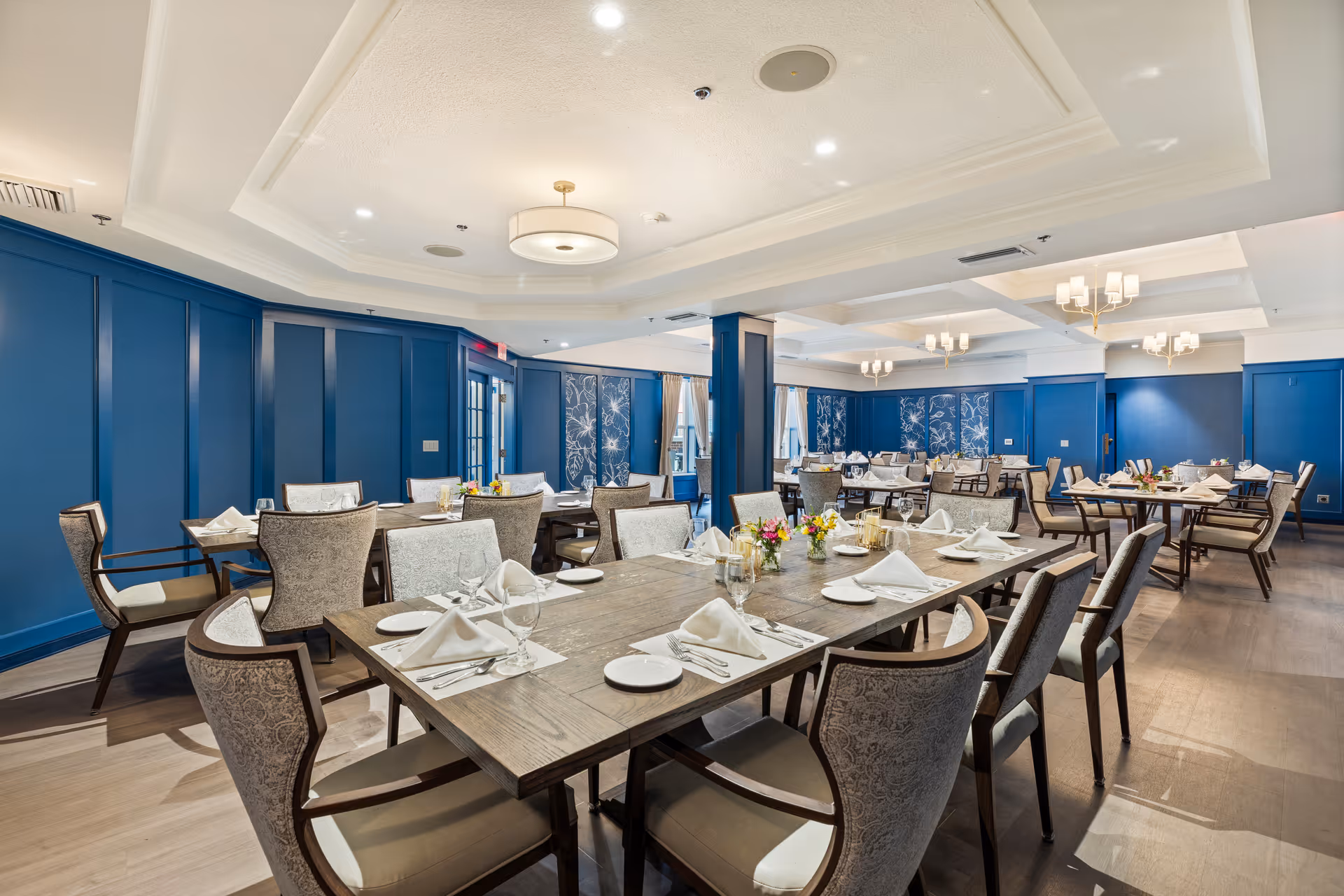 A spacious, elegant dining room with multiple wooden tables set with white napkins, plates, glasses, and silverware. The room features blue paneled walls, decorative wall art, and soft lighting from ceiling fixtures. Comfortable upholstered chairs surround the tables, and small floral centerpieces add a touch of color.