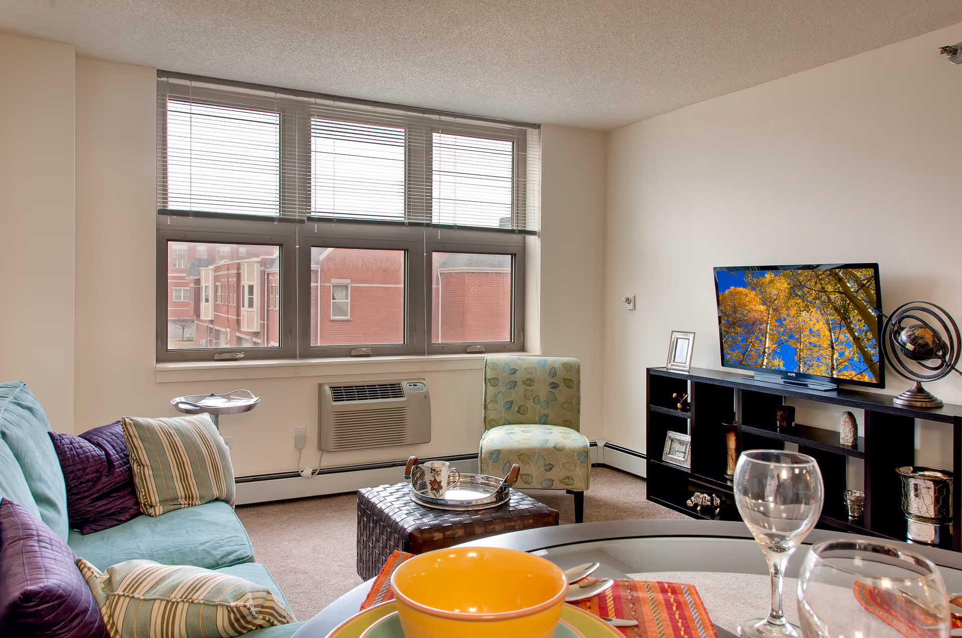 A cozy living room with a teal sofa adorned with striped and purple pillows, a patterned green armchair, a dark woven ottoman with a tray holding a cup and saucer, and a black entertainment unit with a flat-screen TV displaying an image of autumn trees. The room has large windows with blinds, a wall-mounted air conditioner, and a glass dining table set with colorful bowls and wine glasses in the foreground.