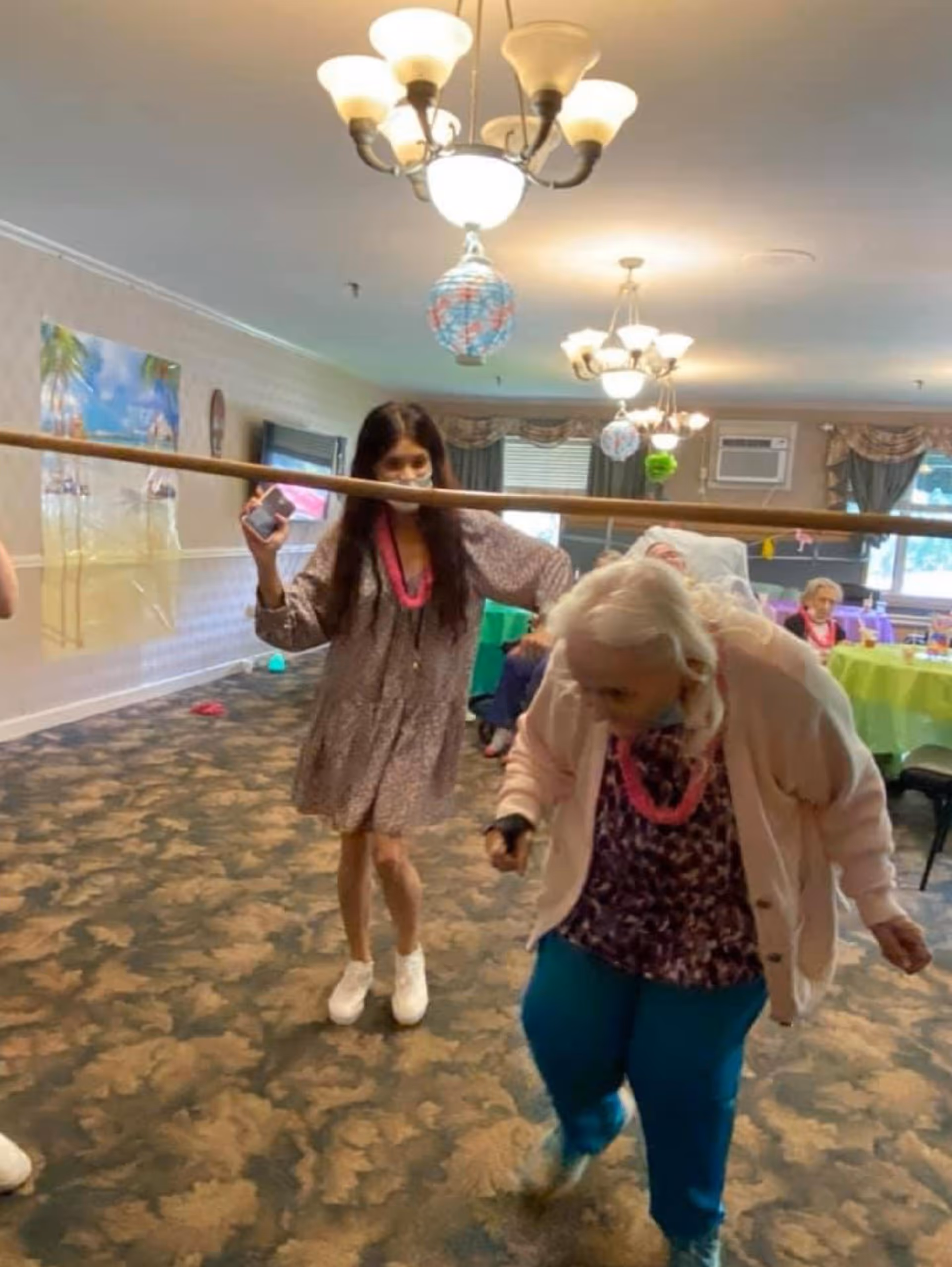 Two women participating in a limbo game inside a decorated room with carpeted floor and chandeliers. One woman is bending backward to pass under a horizontal pole held by the other woman. In the background, there are tables with colorful tablecloths and other seated people.
