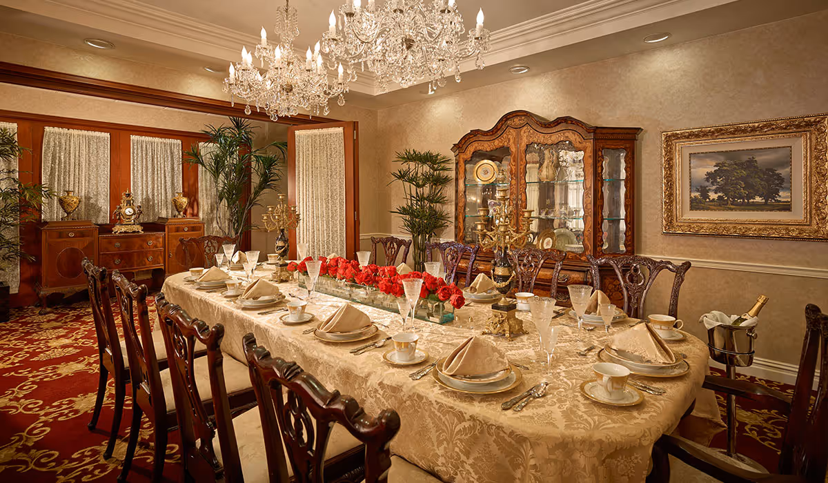 Elegant dining room with a long table covered in a beige patterned tablecloth, set with fine china, folded napkins, glassware, and gold-toned cutlery. The table centerpiece features a rectangular arrangement of red roses. The room has ornate wooden chairs, a crystal chandelier hanging from the ceiling, a wooden china cabinet displaying decorative plates and glassware, and framed artwork on the wall. There are also potted plants and antique-style furniture along the walls.