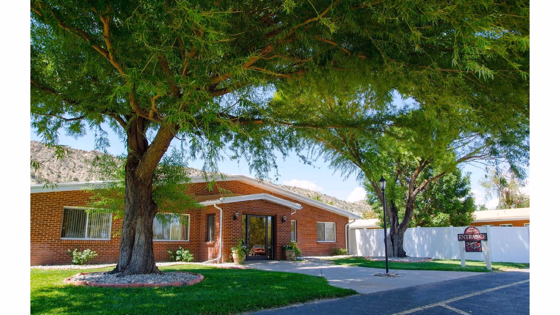 Exterior view of a single-story brick building with a large tree in front, a paved walkway leading to the entrance, and a sign indicating the entrance to Skyline Ridge Nursing and Rehabilitation Center. The area is surrounded by green grass and other trees, with mountains visible in the background under a clear blue sky.