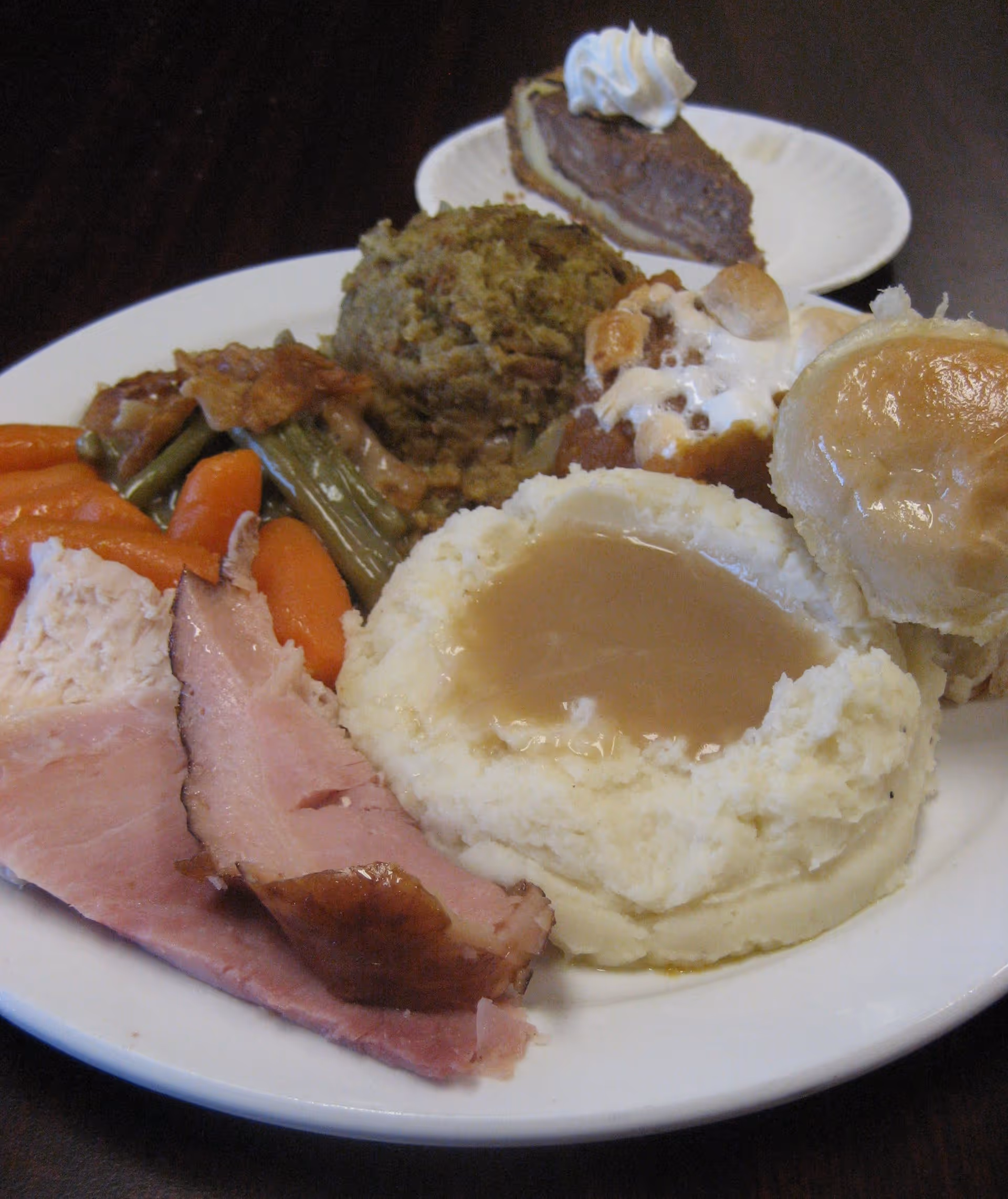 A plate of traditional comfort food including mashed potatoes with gravy, sliced ham, cooked carrots, green beans with bacon, stuffing, a bread roll, and a dessert plate in the background with a slice of pie topped with whipped cream.