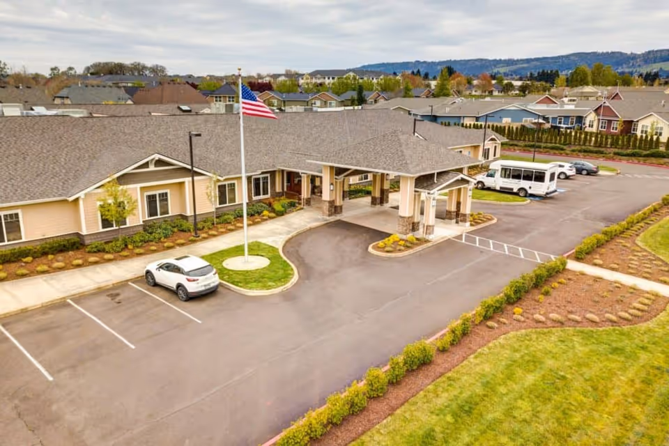 Aerial view of the front entrance of Arbor Oaks Terrace Memory Care Residence, showing a single-story building with a covered drop-off area, an American flag on a flagpole, several parked vehicles, and landscaped grounds with bushes and grass.