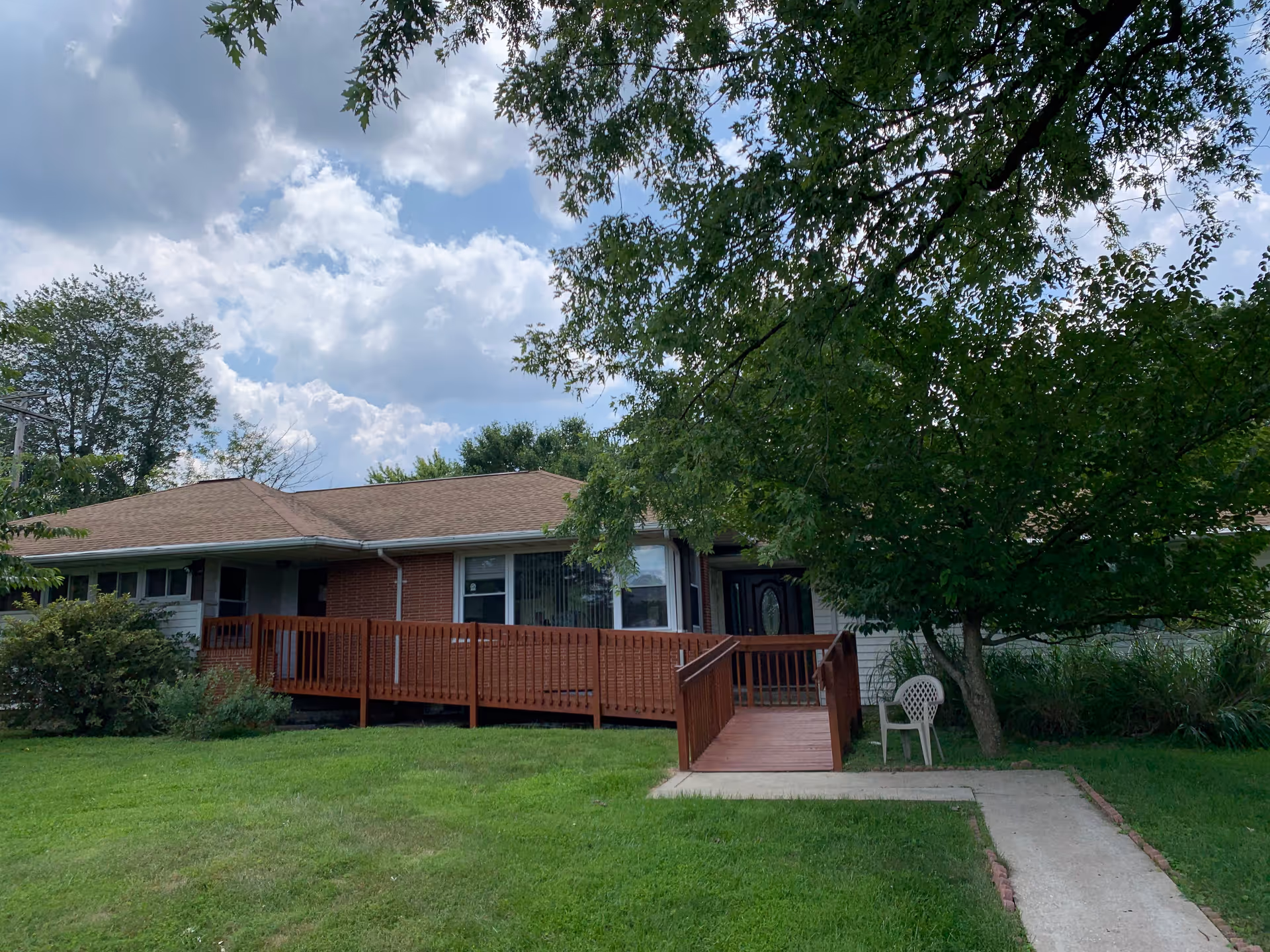 Single-story building with a brown roof and a wooden ramp leading to the entrance, surrounded by green grass and trees under a partly cloudy sky. A white plastic chair is placed near the ramp on the right side.
