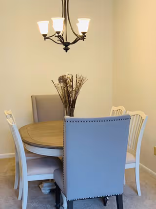 Dining area with a round wooden table, four chairs, a vase of dried branches, and a hanging chandelier.
