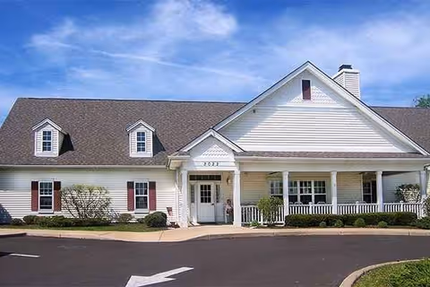 Front exterior view of a single-story building with white siding, a dark shingled roof, and a covered porch with white railings. There are small bushes and landscaping along the front, and a paved driveway with a white directional arrow in front of the entrance.