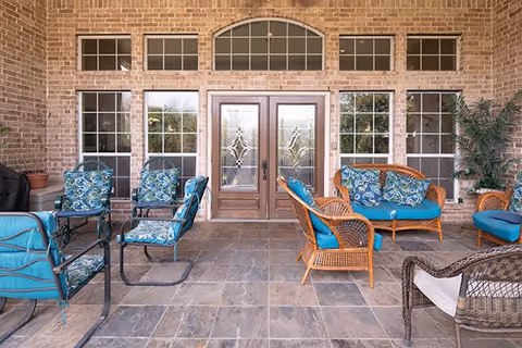 Outdoor patio area with a tiled floor, featuring multiple wicker and metal chairs with blue patterned cushions arranged around a glass-top table. The background shows a brick wall with large windows and double glass doors with decorative designs, and potted plants on either side.
